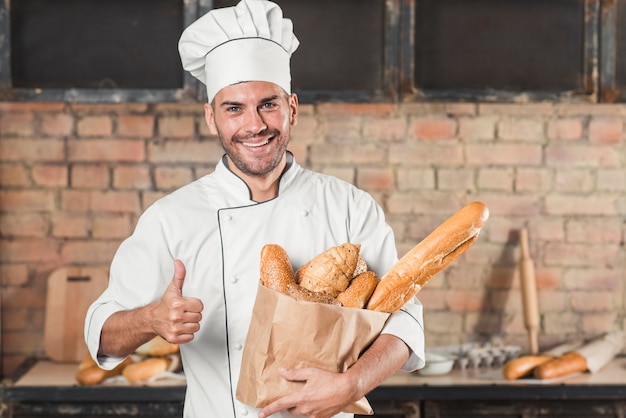 Photo | Smiling young male baker holding loaf of breads in paper bag showing thumb up sign